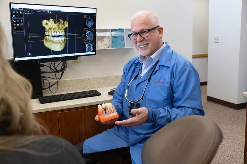 Dr. Robert M. Taylor speaking with a patient while holding a 3D model of dental implants at Taylor and Associates Family and Implant Dentistry in Bay City MI.